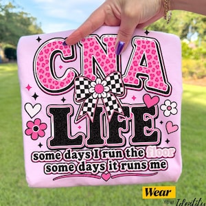 May include: A light pink t-shirt with the words "CNA LIFE" in a bold, playful font. The letters "CNA" are in pink leopard print. The shirt also features a checkered bow and the text "some days I run the floor, some days it runs me."