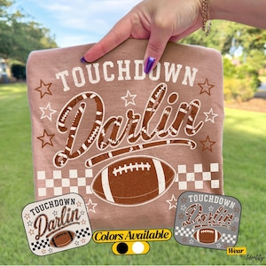 May include: A tan t-shirt featuring the white text TOUCHDOWN and the word Darlin in a brown football-patterned script font. Below is a brown football graphic and a white checkered pattern. The design includes white stars and two small square swatches.
