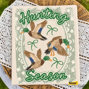 May include: A rectangular canvas print featuring the text Hunting Season in dark green script. The design includes three flying mallard ducks with brown, green, and blue plumage, accented by small green bows against a light background.