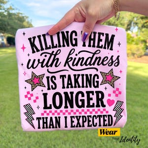 May include: A light pink t-shirt featuring black text that reads "KILLING THEM with kindness IS TAKING LONGER THAN I EXPECTED." The design includes black and pink star graphics with leopard print, pink checkerboard patterns, and black lightning bolts.