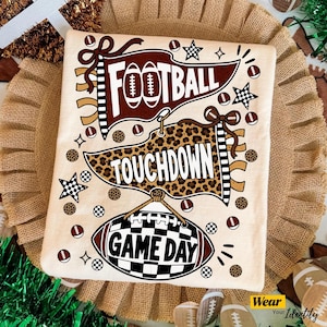 May include: A cream-coloured t-shirt featuring a graphic print with three pennants and football motifs. The top pennant is maroon with the text FOOTBALL, the middle is leopard print with TOUCHDOWN, and the bottom is a football with GAME DAY.