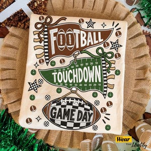 Puede incluir: Una camiseta color crema con un diseño gráfico de tres banderines y un balón de fútbol americano. El banderín marrón superior dice FOOTBALL, el verde central dice TOUCHDOWN y el gráfico inferior con forma de balón dice GAME DAY.