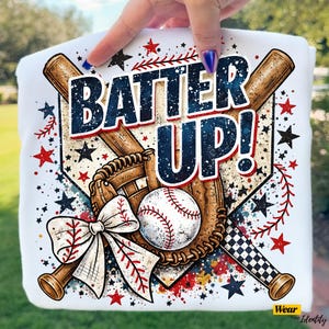 Puede incluir: Camiseta blanca con un diseño de béisbol. El diseño presenta una pelota de béisbol, un guante, bates y las palabras "BATTER UP!" en azul marino y blanco. Estrellas rojas y negras y un lazo blanco completan el diseño.