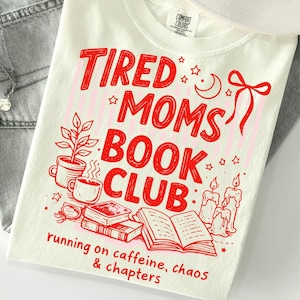 Op de afbeelding: Een crèmekleurig t-shirt met rode grafische tekst die TIRED MOMS BOOK CLUB en running on caffeine, chaos & chapters leest. Het ontwerp bevat illustraties van een kamerplant, koffiemokken, gestapelde boeken, een open boek en kaarsen.