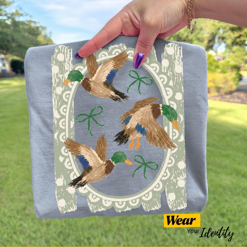 May include: A light blue t-shirt featuring a graphic print of three flying mallard ducks in brown, green, and blue. The ducks are framed by a decorative sage green oval border with white scalloped edges and small green ribbon accents.