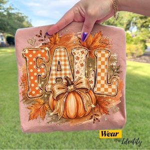 May include: A light pink t-shirt featuring a graphic print with the word FALL in large, patterned orange letters. The design includes a central orange pumpkin with a large bow, surrounded by autumn leaves, small hearts, and sparkling star accents.