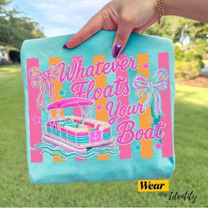 May include: A light blue t-shirt featuring a graphic print of a pink and teal pontoon boat on water, surrounded by pink bows and stars. The text "Whatever Floats Your Boat" is printed in a pink script font over vertical orange and pink stripes.