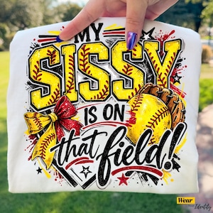 May include: White t-shirt with a baseball-themed graphic. The design features the text "MY SISSY IS ON that field!" in yellow and black, along with a baseball, glove, and a red and yellow bow.