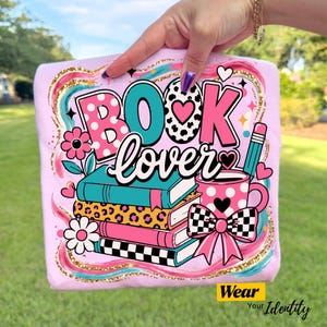 May include: A light pink t-shirt featuring a graphic print with the text BOOK lover. The design includes a stack of four books, a pink polka-dot mug with a pencil, a checkered bow, and flowers, all surrounded by colorful swirling brush strokes.
