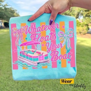 May include: A light blue t-shirt with the phrase "Whatever Floats Your Boat" in pink and teal script. The design includes a pink and teal pontoon boat, bows, and stars. The shirt has orange and pink vertical stripes.