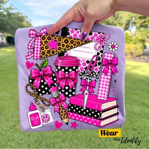 May include: A light purple t-shirt featuring a graphic print of school supplies including a leopard print pennant, a polka dot apple, a coffee cup, a pencil, scissors, and books, all decorated with bright pink bows and gingham patterns.