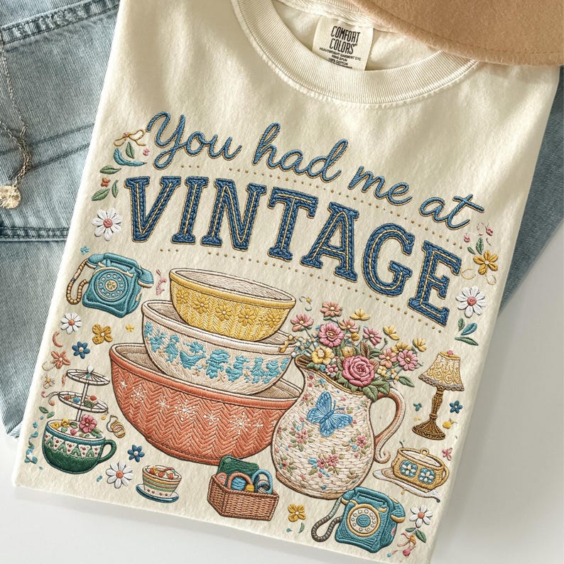 May include: A cream-colored t-shirt featuring the text You had me at VINTAGE in blue embroidery-style font. The graphic includes illustrations of stacked mixing bowls, a floral pitcher, rotary telephones, a lamp, and small flowers.