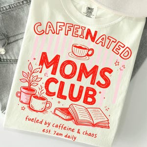 Puede incluir: Una camiseta de color crema con texto rojo que dice CAFFEINATED MOMS CLUB y fueled by caffeine & chaos est. 7am daily. El gráfico incluye ilustraciones de una taza de café, una planta en maceta y un libro abierto sobre rayas rosas.