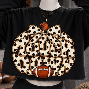May include: A black long-sleeve cropped sweatshirt featuring a large graphic of a pumpkin with a polka dot pattern and a bow. The text "TOUCH DOWN" is printed in the center of the pumpkin, accompanied by a brown football graphic at the bottom.