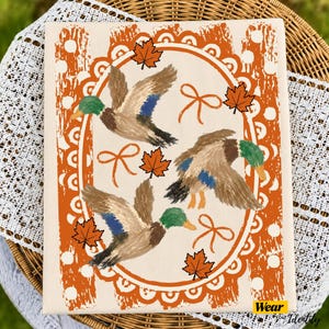 Pode incluir: Uma tela retangular com três patos-reais em voo, rodeados por folhas de ácer cor de laranja e laços desenhados com linhas simples. A moldura oval central tem uma borda recortada sobre um fundo de riscas verticais cor de laranja e brancas.