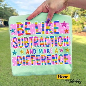 May include: A light green t-shirt featuring the text BE LIKE SUBTRACTION AND MAKE A DIFFERENCE in multi-colored, striped letters. The design includes pink rectangular backgrounds with white polka dots and small, colorful starfish illustrations.