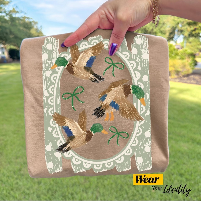 May include: A tan t-shirt featuring a graphic print of three mallard ducks in flight, arranged within a light green scalloped oval frame. The design includes small green ribbon accents and a textured, distressed border pattern on the sides.