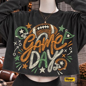 May include: A charcoal grey cropped sweatshirt featuring a graphic print with the text "GAME DAY" in orange and white. The design includes illustrations of a brown football, two football helmets, stars, and various play-calling symbols.