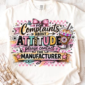 Pode incluir: Camiseta creme com um gráfico colorido que diz "For Complaints About Attitude Please Contact The Manufacturer". O design inclui um laço rosa, uma borboleta e uma xícara de café.