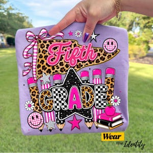 May include: A light purple t-shirt featuring a graphic with the text Fifth Grade. The design includes a leopard print pennant, pink and black pencils, a black star, a pink apple on books, smiley faces, and flowers in shades of pink and white.