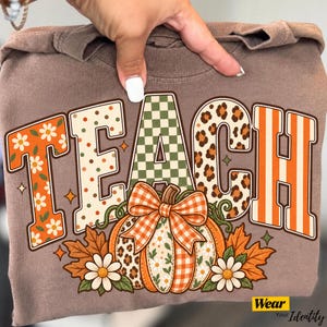 Könnte beinhalten: Ein braunes T-Shirt mit dem Wort TEACH in großen Blockbuchstaben, die jeweils ein einzigartiges Muster aus orangefarbenen Blumen, Punkten, grünen Karos, Leopardenmuster und orangefarbenen Streifen aufweisen, mit Kürbis und Herbstlaub.