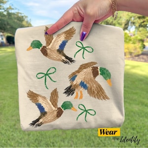May include: A beige t-shirt featuring a printed design of three mallard ducks in flight, each with brown, green, and blue plumage, interspersed with three simple green line-drawn bows. The bottom corner displays a yellow tag with the text Wear Your Identity.