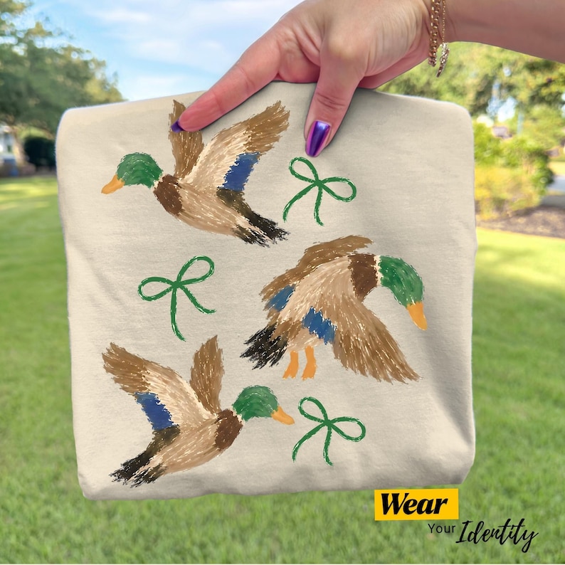 May include: A beige t-shirt featuring a printed design of three mallard ducks in flight, each with brown, green, and blue plumage, interspersed with three simple green line-drawn bows. The bottom corner displays a yellow tag with the text Wear Your Identity.