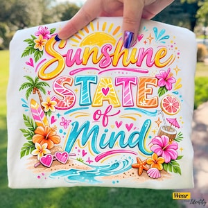 Puede incluir: Camiseta blanca con un diseño gráfico colorido. El diseño presenta las palabras "Sunshine State of Mind" en una fuente lúdica, junto con ilustraciones de un sol, flores, una tabla de surf y una bebida tropical. La camiseta es sostenida por una persona.