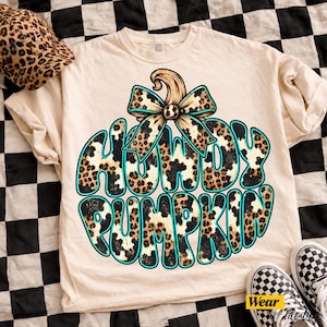 May include: A cream-colored t-shirt featuring a graphic of a pumpkin shape filled with a leopard print pattern. The text "HOWDY PUMPKIN" is written in a bold, bubble font with a leopard print fill and a bright turquoise outline. A large leopard print bow sits atop the pumpkin graphic.