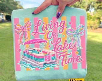 Könnte beinhalten: Hellblaues T-Shirt mit rosa-orangenem Streifendesign. Das Shirt zeigt eine Grafik eines Pontonboots und die Worte "Living On Lake Time" in rosa Schrift. Zwei Schleifen und Sterne sind ebenfalls enthalten.