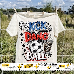 May include: Cream-colored t-shirt with a soccer-themed graphic. The design features the text "Kick the Dang Ball" in blue and red, a soccer ball, stars, and a drink. The shirt is hanging on a clothesline.