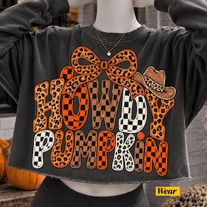 May include: A charcoal gray cropped sweatshirt featuring a graphic print with the text HOWDY PUMPKIN. The design includes a large leopard print bow, a cowboy hat, and letters filled with orange and black checkered patterns and leopard print spots.