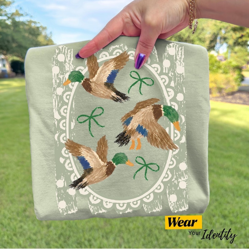 May include: A sage green t-shirt featuring a white scalloped oval frame containing three flying mallard ducks with brown, green, and blue plumage, accented by three small green bows. The design is printed on the front of the fabric.
