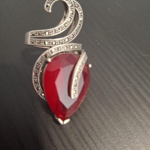 Puede incluir: Un anillo plateado con una gran piedra preciosa roja facetada en forma de lágrima. El anillo presenta un diseño decorativo en espiral con pequeñas piedras incrustadas. La banda y el diseño en espiral son de color plateado.
