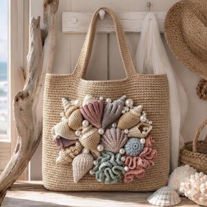 May include: A beige crocheted tote bag featuring a central cluster of three-dimensional crocheted seashells and coral-like shapes in shades of pink, blue, gray, and tan, accented with scattered white faux pearls attached to the front surface.