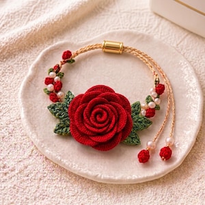 May include: A bracelet featuring a large crocheted red rose centerpiece with green crocheted leaves, accented by small white pearls and tiny red crocheted buds on a braided tan cord, finished with a gold-toned cylindrical metal clasp.