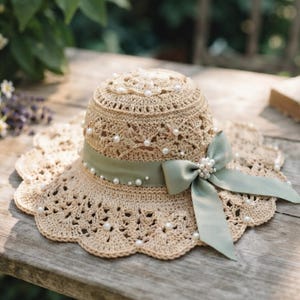 May include: A beige crocheted sun hat featuring an intricate openwork pattern, adorned with scattered white faux pearls throughout the crown and scalloped brim. A sage green satin ribbon is tied in a bow, accented with a cluster of pearls.
