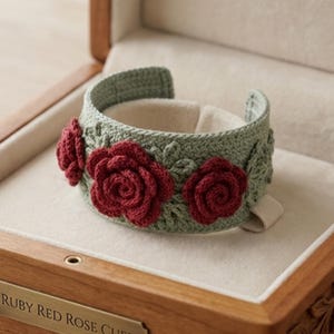 May include: A sage green crochet cuff bracelet featuring three dimensional dark red rose appliqués and intricate leaf detailing. The cuff is open-ended and rests inside a wooden box with a plaque reading RUBY RED ROSE CUFF.