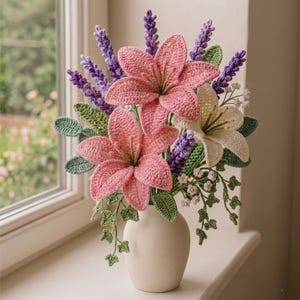May include: A crochet flower arrangement in a matte white vase featuring two large pink lilies, one cream lily, purple lavender sprigs, small white blossoms, and green leaves. The handcrafted textile bouquet displays intricate yarn textures.