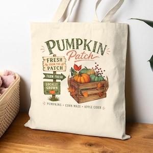 May include: A cream-colored canvas tote bag featuring a graphic print of a wooden crate filled with pumpkins. The text reads PUMPKIN Patch, FRESH FROM THE PATCH, FARM FRESH, LOCALLY GROWN, PUMPKINS, CORN MAZE, and APPLE CIDER with a signpost illustration.