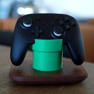 May include: A black wireless video game controller resting on a bright green cylindrical stand, which is mounted on a square, dark brown wooden base. The controller features two analog sticks, a directional pad, and four lettered action buttons.