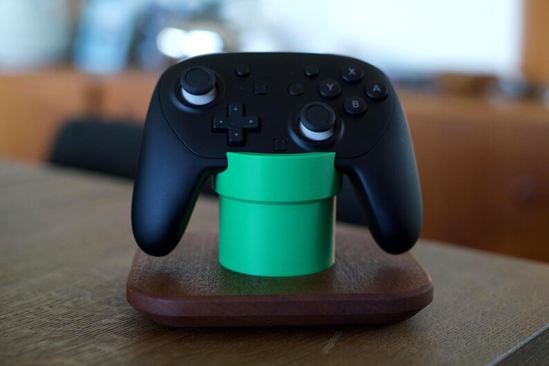 May include: A black wireless video game controller resting on a bright green cylindrical stand, which is mounted on a square, dark brown wooden base. The controller features two analog sticks, a directional pad, and four lettered action buttons.