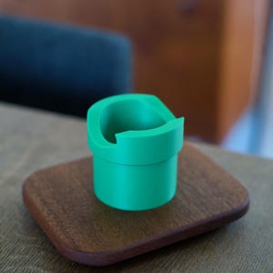 May include: A bright green, cylindrical plastic holder with a wide, open top and a distinct notched rim. The object features a smooth, matte finish and a hollow interior, designed with a tiered structure that narrows slightly toward the base.