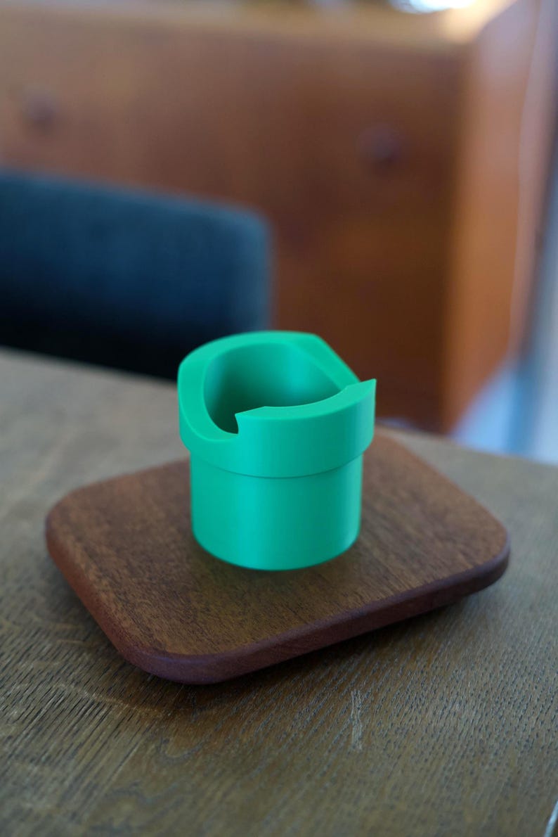 May include: A bright green, cylindrical plastic holder with a wide, open top and a distinct notched rim. The object features a smooth, matte finish and a hollow interior, designed with a tiered structure that narrows slightly toward the base.