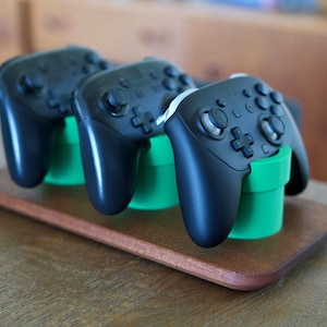 May include: Three black wireless video game controllers are positioned upright in three bright green cylindrical stands. These stands are mounted on a rectangular wooden base with rounded edges, providing a stable display for the controllers.