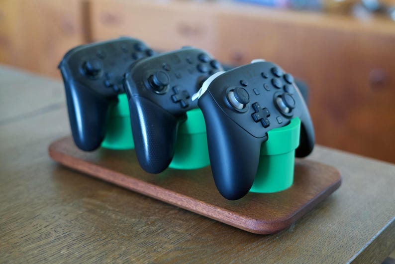 May include: Three black wireless video game controllers are positioned upright in three bright green cylindrical stands. These stands are mounted on a rectangular wooden base with rounded edges, providing a stable display for the controllers.