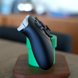 May include: A black and white video game controller resting in a bright green cylindrical stand. The stand is mounted on a smooth, rectangular wooden base with rounded corners, providing a stable platform for the gaming accessory.