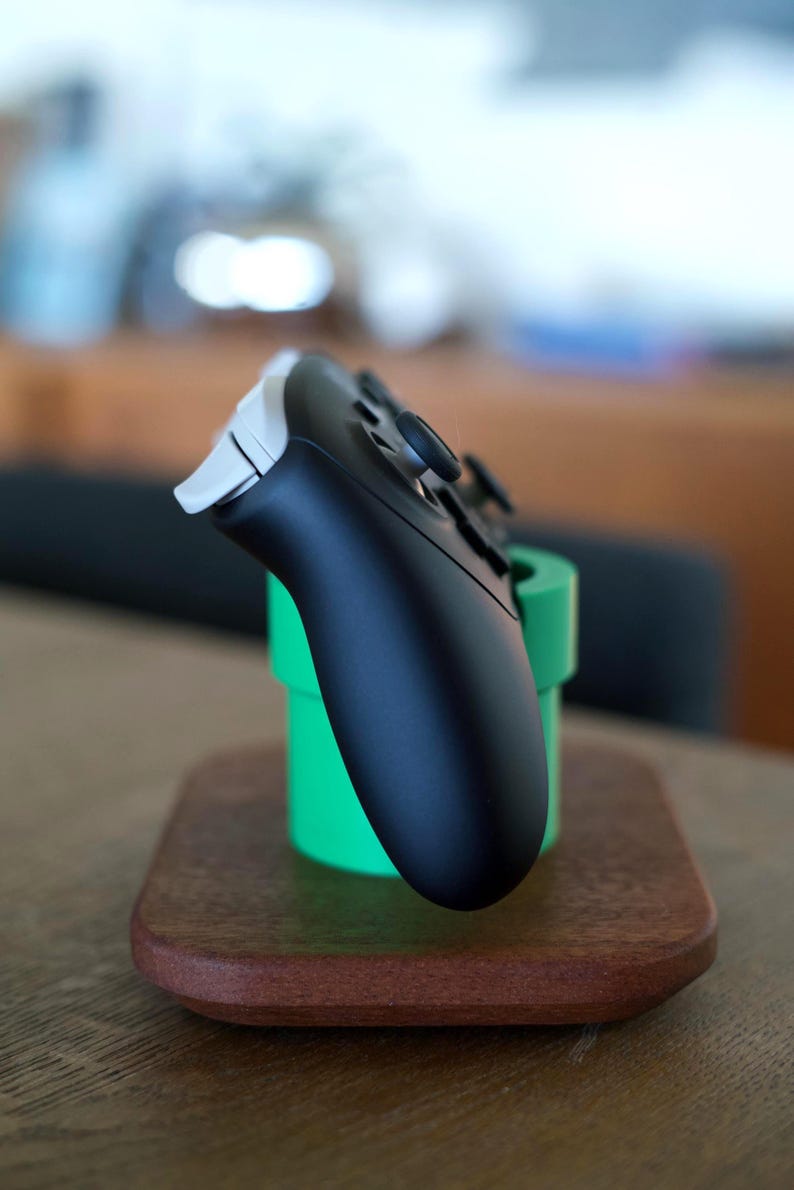 May include: A black and white video game controller resting in a bright green cylindrical stand. The stand is mounted on a smooth, rectangular wooden base with rounded corners, providing a stable platform for the gaming accessory.