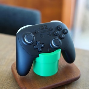 May include: A black Nintendo Switch Pro controller resting on a bright green cylindrical stand, which is mounted on a rectangular wooden base. The controller features standard buttons, dual analog sticks, and the Nintendo Switch logo printed in white.