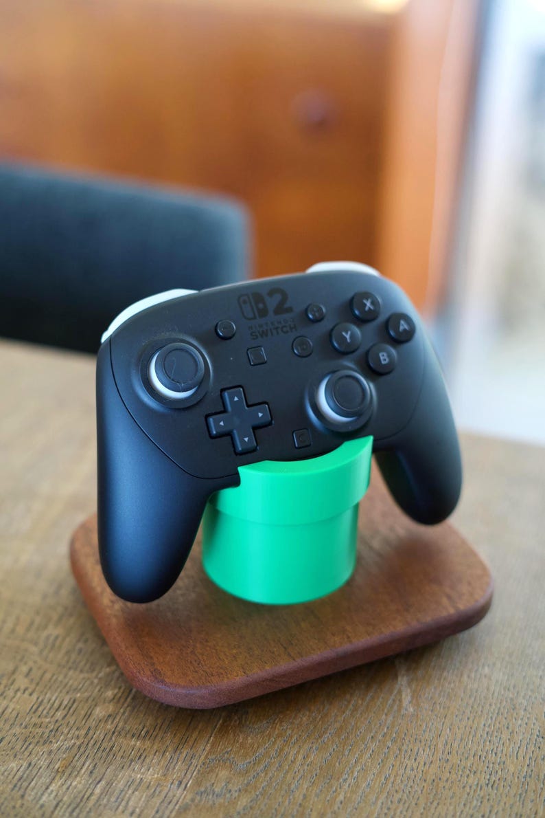 May include: A black Nintendo Switch Pro controller resting on a bright green cylindrical stand, which is mounted on a rectangular wooden base. The controller features standard buttons, dual analog sticks, and the Nintendo Switch logo printed in white.